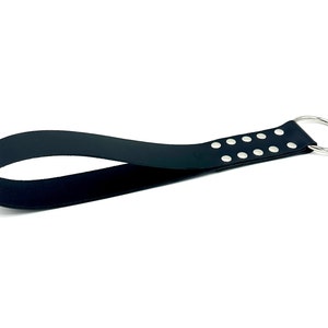 May include: A black leather wrist strap with a silver metal ring and silver rivets. The strap is curved and appears to be made of a single piece of leather. The background is white.