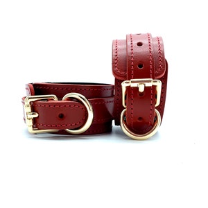 May include: A pair of red leather wrist cuffs with gold metal buckles and D-rings.