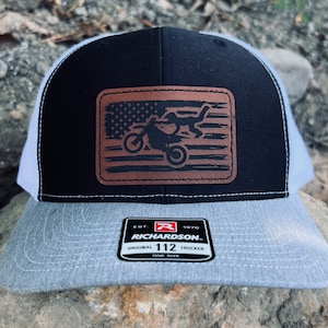 May include: A black and white trucker hat with a brown leather patch featuring an American flag and a silhouette of a dirt bike. The patch has the text "EST. 1970 RICHARDSON. ORIGINAL 112 TRUCKER ONE SIZE" on it.