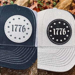 May include: Two trucker hats with a black and white leather patch that says "1776" surrounded by stars. The hat on the left is black with a gray mesh back. The hat on the right is gray with a black mesh back.