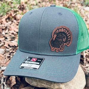 Puede incluir: Una gorra de camionero gris oscuro y verde con un parche de cuero marrón de un pavo en la parte delantera. La gorra es ajustable y tiene el logotipo de Richardson en el lateral.