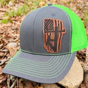 Può includere: Cappellino da camionista grigio e verde neon con una toppa in pelle marrone. La toppa presenta un disegno della bandiera americana e la silhouette di un elettricista su un palo della luce. Il cappellino ha una visiera curva con cuciture verde neon.