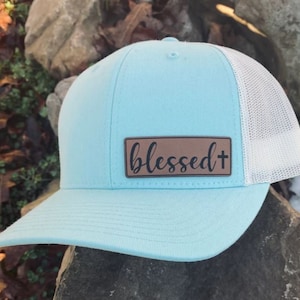 May include: A light blue and white trucker hat with a brown leather patch that says "blessed+"