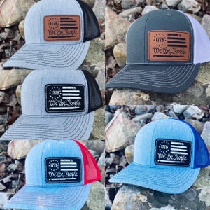 Puede incluir: Cinco gorras de camionero con una malla negra en la parte posterior y un parche de cuero marrón con una bandera estadounidense blanca y el texto "1776: We Be People". Las gorras son de diferentes colores: gris, gris oscuro, azul claro, azul y azul claro con una malla roja en la parte posterior.
