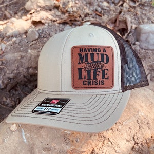 May include: A beige and black trucker hat with a brown leather patch that says "Having a Mud Life Crisis". The patch has a black tire tread design.