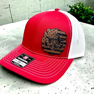 May include: Red and white trucker hat with a brown leather patch featuring a firefighter helmet, axe, and American flag. The patch says "FD" on the helmet. The hat is labeled "Richardson 112 Original Trucker Adjustable."