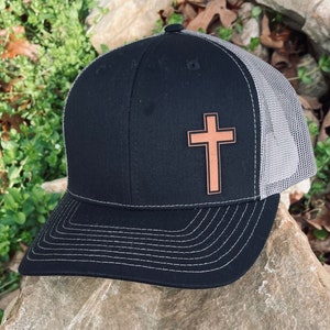 May include: A black and gray trucker hat with a brown leather cross patch on the front.