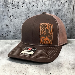 May include: A brown and tan trucker hat with a brown leather patch featuring a silhouette of a soldier saluting in front of an American flag.