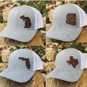 May include: Four grey and white trucker hats with different state outlines in brown leather. The states are Michigan, Arizona, Florida, and Texas.