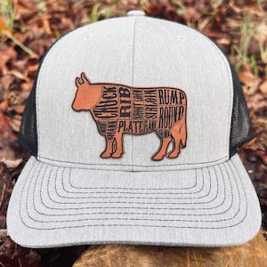 Peut inclure: Un chapeau de camionneur gris et noir avec un patch en cuir brun représentant une vache avec les différentes coupes de viande étiquetées. Le patch comprend les mots "Chuck", "Rib", "Short loin", "Sirloin", "Rump", "Round", "Plate", "Flank", et "Shank".