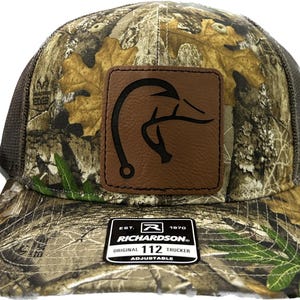 May include: A camouflage trucker hat featuring a brown leather patch on the front with a black graphic of a fish hook and deer antler. The hat has a mesh back and a black sticker on the brim that reads EST. 1970 RICHARDSON ORIGINAL 112 TRUCKER ADJUSTABLE.