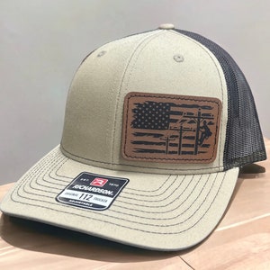 May include: A beige and black trucker hat with a brown leather patch featuring an American flag and a silhouette of a lineman working on a power line. The patch is stitched onto the front of the hat. The hat is adjustable and has the text "Richardson 112 Trucker Adjustable" on the back.