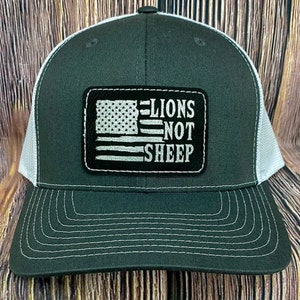 May include: A black and white trucker hat with a white mesh back. The hat has a patch on the front with a gray and white American flag and the text "LIONS NOT SHEEP".