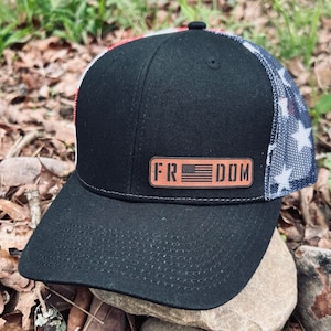 May include: Black trucker hat with a red, white, and blue American flag mesh back. The hat has a brown leather patch with the word "FREEDOM" and a small American flag.