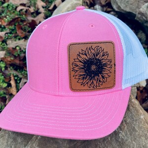 May include: Pink trucker hat with a white mesh back and a curved brim. A brown leather patch on the front features a black sunflower design. The hat is made of a durable fabric and has a snapback closure.