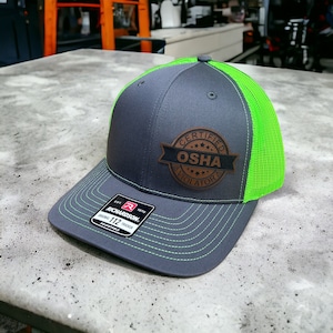 Könnte beinhalten: Eine graue und grüne Trucker-Kappe mit einem braunen Leder-Patch, auf dem "Certified OSHA Violator" steht.