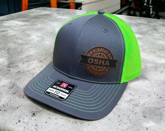 Könnte beinhalten: Eine graue und grüne Trucker-Kappe mit einem braunen Leder-Patch, auf dem "Certified OSHA Violator" steht.
