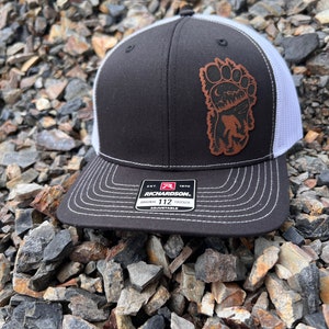 Op de afbeelding: Zwarte en witte truckerpet met een Bigfoot-patch. De pet heeft een zwart voorpaneel, een witte mesh achterkant en een gebogen klep met witte stiksels. De patch toont een Bigfoot-silhouet met een berg- en maanlandschap.