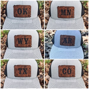May include: Five trucker hats with different state abbreviations on them. The hats are all gray with black mesh backs. The state abbreviations are OK, MN, WY, AR, and CO. The letters are black and the background is brown leather with a floral design.