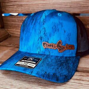 May include: Blue trucker hat with a black mesh back. The hat features a leather patch with the words "FISHING LIFE" and a fish graphic. The hat is adjustable and has a Richardson 112 tag.