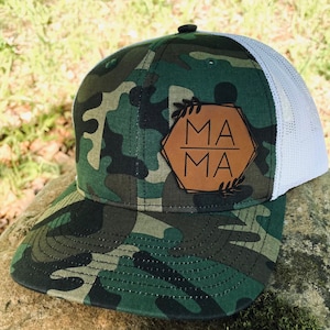 Może przedstawiać: Czapka bejsbolowa w kamuflażu z białym tyłem z siatki. Przód czapki zdobi brązowa skórzana naszywka z napisem "MAMA" w czarnych literach, obramowana geometrycznym wzorem i akcentami liści. Czapka spoczywa na kamieniu.