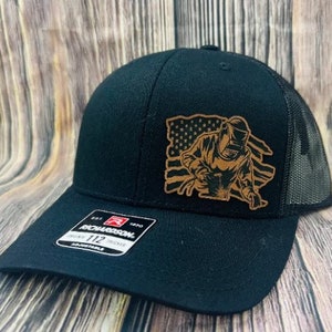 Può includere: Cappellino da camionista nero con una toppa in pelle marrone che raffigura un saldatore al lavoro davanti a una bandiera americana. Il cappello è realizzato da Richardson e ha una fibbia regolabile.