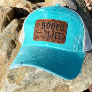 May include: A turquoise trucker hat with a distressed look. The hat features a brown leather patch with the words "RODEO LIFE" in black lettering. The hat has a white mesh back and a curved brim.