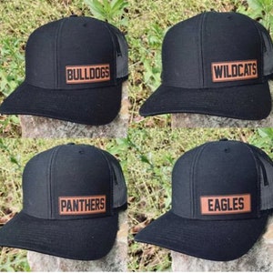 May include: Four black baseball caps with mesh backs and curved brims. Each cap has a rectangular patch with a team name in orange lettering: Bulldogs, Wildcats, Panthers, and Eagles. The caps are displayed on a natural, textured surface.
