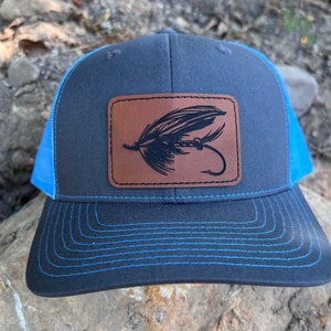 May include: A black and blue trucker hat with a brown leather patch featuring a black silhouette of a fly fishing lure.