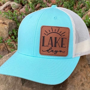 May include: A light blue trucker hat with a white mesh back. The hat has a brown leather patch with a sun and the text "LAKE days" in black.