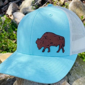 May include: A light blue trucker hat with a white mesh back. The hat features a brown leather buffalo silhouette.