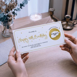 Könnte beinhalten: Eine weiße und goldene Geburtstagskarte mit dem Text "Happy 18th Birthday" und "To Amelia". Die Karte hat einen Rubbelbereich, der "You are going to Paris!" enthüllt. Die Karte wird vor einem braunen Umschlag gehalten.