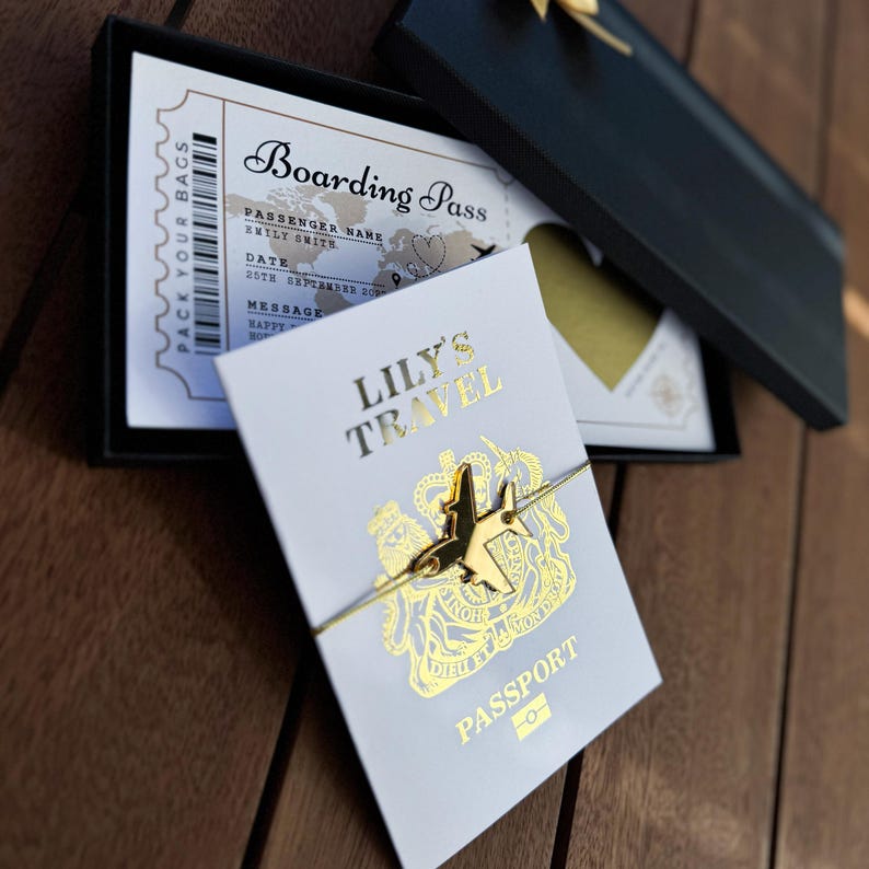 May include: A gold-toned airplane charm bracelet on a white card with gold foil text reading LILY'S TRAVEL PASSPORT, placed over a black gift box containing a printed boarding pass with the text PACK YOUR BAGS and BOARDING PASS.