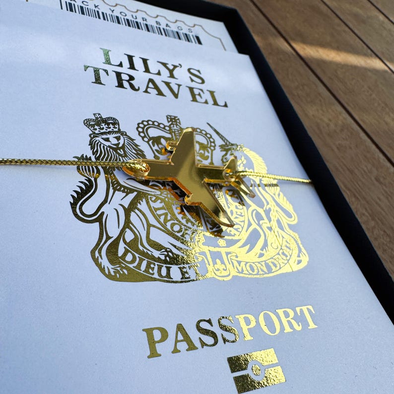 May include: A gold-colored airplane charm attached to a thin gold cord, resting on a white card with gold foil text reading LILY'S TRAVEL and PASSPORT, featuring a gold royal coat of arms emblem with a lion and unicorn design on the front.