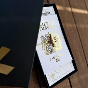 May include: A gold-toned airplane charm bracelet resting on a white card printed with gold foil text reading LILY'S TRAVEL PASSPORT and a royal crest, all housed inside a black gift box with a gold ribbon accent on the lid.