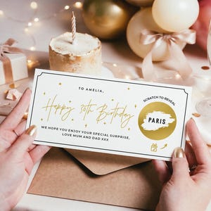 Könnte beinhalten: Eine Geburtstagskarte mit goldfarbenem Schriftzug "Happy 30th Birthday" und "To Amelia". Die Karte hat einen Rubbelbereich mit der Aufschrift "Paris". Die Karte wird über einem braunen Umschlag gehalten.