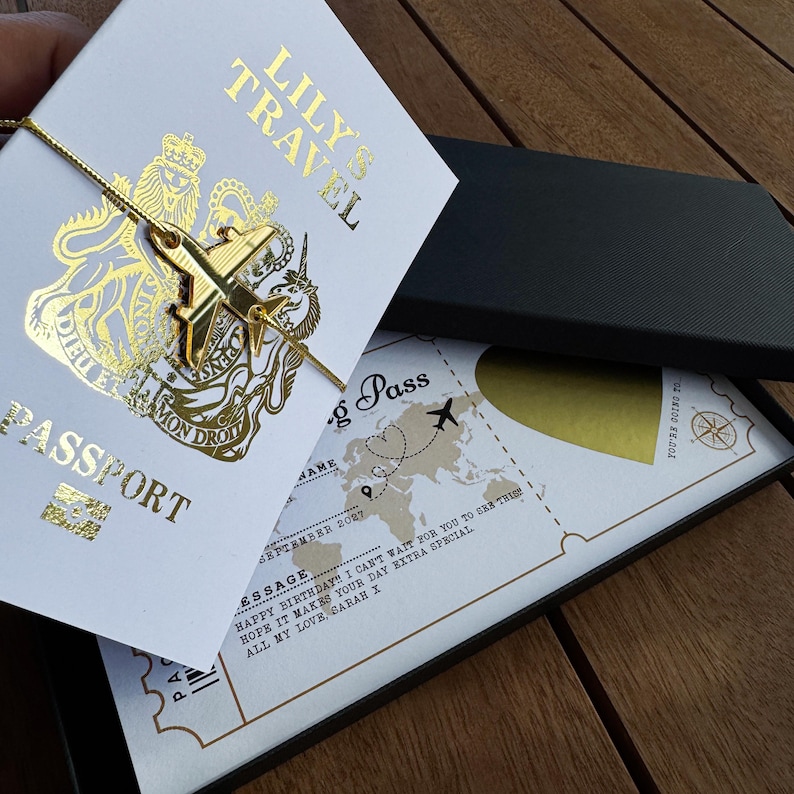 May include: A white passport-style card with gold foil text reading LILY'S TRAVEL and PASSPORT, featuring a gold airplane charm tied with a gold string. Beneath it lies a boarding pass with a world map, a gold scratch-off heart, and printed text.
