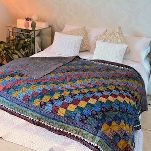 May include: A colorful patchwork quilt with a gray throw blanket on top. The quilt is made of squares of different colors and patterns, including blue, green, yellow, and red. The quilt is on a white bed with white pillows.