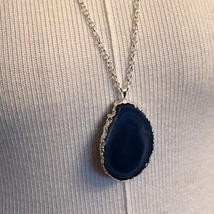May include: A necklace featuring a dark blue, oval-shaped agate slice pendant with a metallic silver-toned edge. The pendant is suspended from a delicate silver-toned rolo chain, showcasing a smooth, polished stone surface with natural banding.