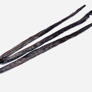 Puede incluir: Tres vainas de vainilla largas y de color marrón oscuro sobre un fondo blanco.