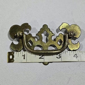 May include: A brass-toned drawer pull with an ornate design. The pull has a curved handle and decorative flourishes on each end. The pull measures approximately 4 inches in length.