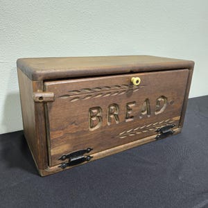 May include: A wooden bread box with a hinged door and a dark brown finish. The word "BREAD" is carved into the front, with decorative wheat stalks. The box has black metal hinges and a yellow knob.