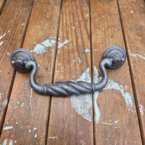 May include: A dark gray metal drawer pull with a twisted handle design. The pull has two circular mounting plates on each end with a curved handle. The hardware is made of cast iron.