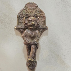 May include: A bronze-colored, decorative door knocker featuring a stylized figure with a mustache and a decorative arched top. The figure is in a seated position with one leg crossed over the other. The knocker has a pointed bottom.