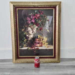 May include: A framed still life print featuring a cherub, flowers, and grapes. The artwork is in a gold frame with a burgundy inner border. The print depicts a classical arrangement of flowers, grapes, and a cherub statue. A Coca-Cola can is in the foreground.