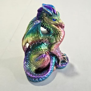 Peut inclure: Figurine de dragon multicolore avec une finition irisée arc-en-ciel. Le dragon a des yeux bleus, une crête violette et des ailes aux couleurs variées. La sculpture est détaillée avec des écailles et est assise enroulée.