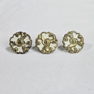 Könnte beinhalten: Drei antike Schrankknöpfe mit floralem Design. Jeder Knopf hat eine weiße Mitte mit goldfarbenem Filigran und einen dunkelbronzenen Außenrand. Die Knöpfe sind rund und haben eine klassische, vintage Ästhetik.