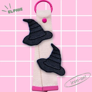 Könnte beinhalten: Zwei schwarze Hexenhut-Aufnäher auf einem cremefarbenen Riemen mit einem rosa Knopf und Ring. Das Wort "ELPHIE" befindet sich oben links und "iron-on!" unten rechts.