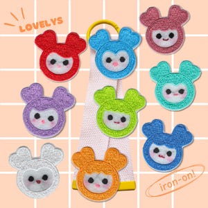 May include: A collection of nine circular embroidered patches featuring stylized faces with heart-shaped ears in red, blue, pink, teal, purple, lime green, white, orange, and dark blue, arranged around a central light pink fabric strap.