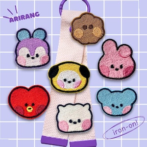 Könnte beinhalten: Eine Sammlung bunter, aufbügelbarer Aufnäher in verschiedenen Formen, darunter ein Herz, ein Bär und ein Hase. Die Aufnäher sind an einem hellrosa Band mit einem lila Ring befestigt. Das Wort "ARIRANG" ist oben aufgedruckt.